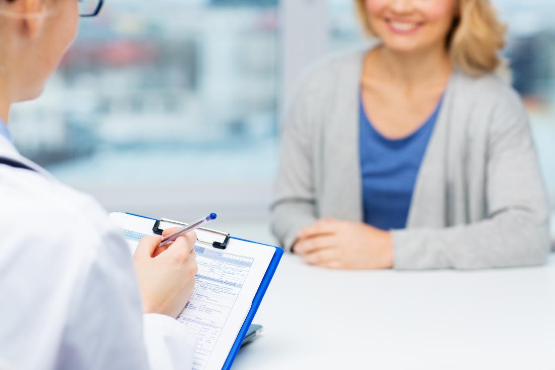 Doctor with clipboard in foreground sitting across from smiling woman.