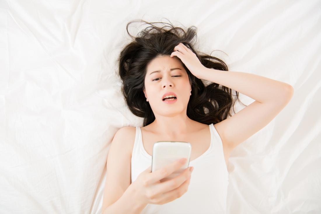 Woman with head pain laying on bed and looking at phone