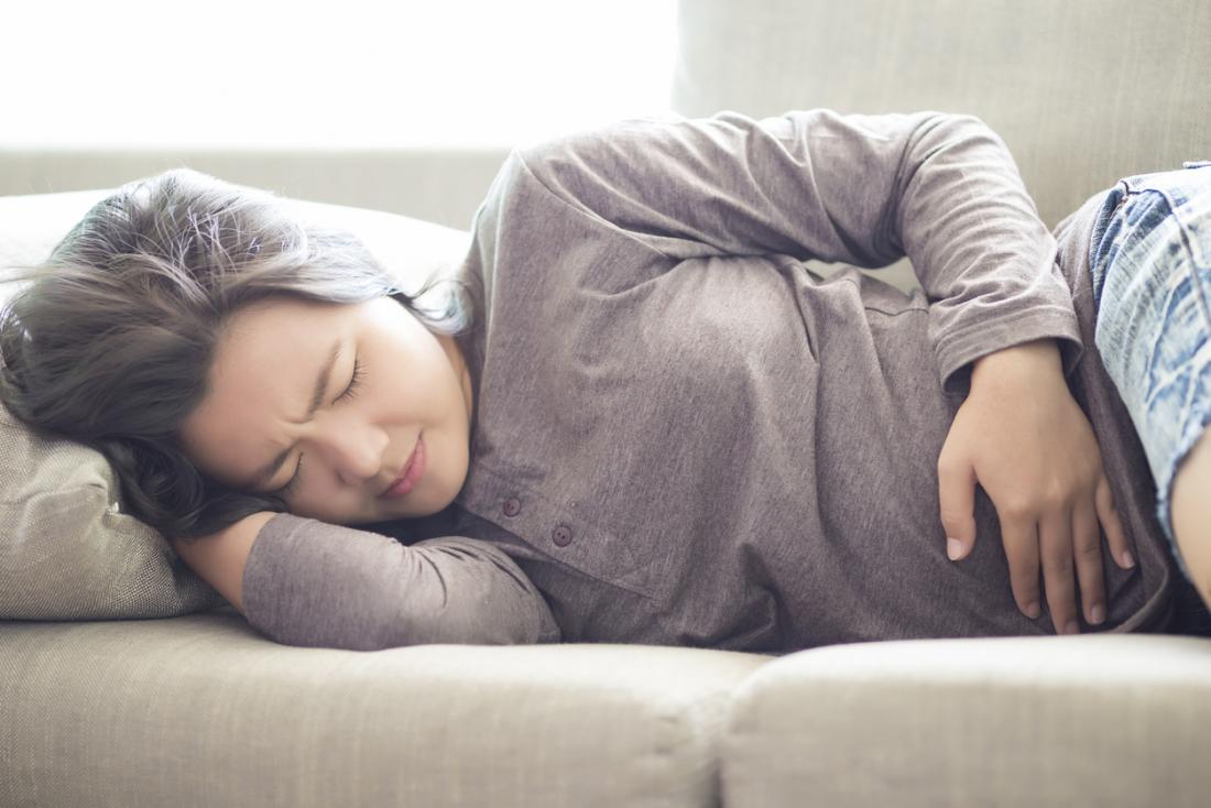 Woman lying on her side on a sofa, holding her stomach in pain, experiencing period cramps and bloating.