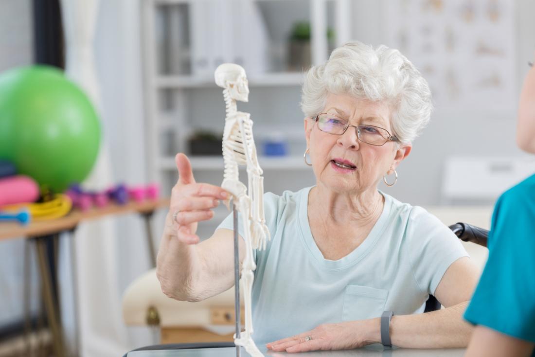 Senior woman talking to osteopath and chiropractor, pointing to model of skeleton to describe her pain.