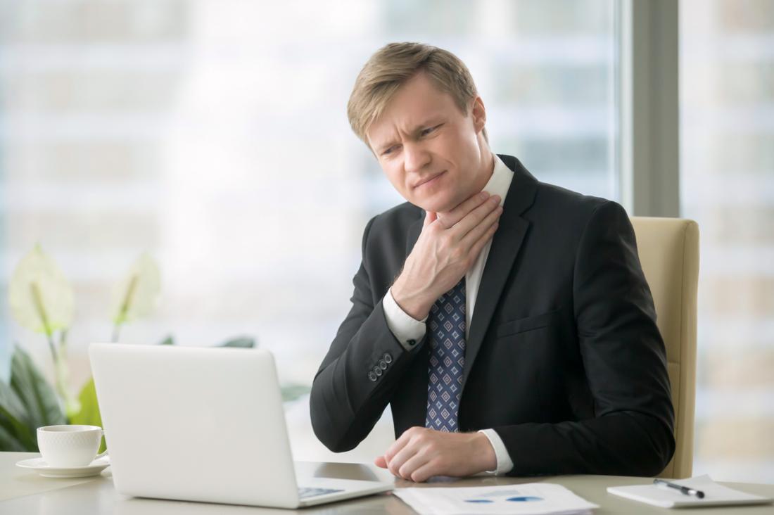 Man in business suit at desk in front of laptop, holding his sore throat in pain.
