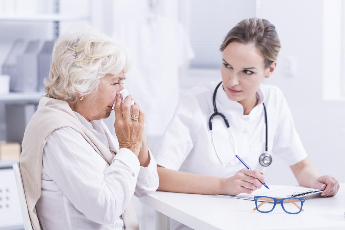 Senior woman blowing her nose speaking to young doctor about allergies and hayfever.