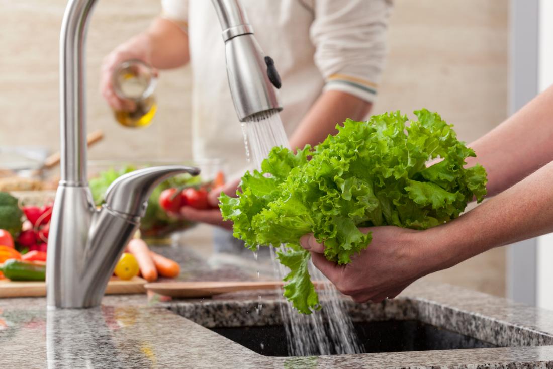 Person preparing vegetables, washing curly kale leaves under running tap water in kitchen.