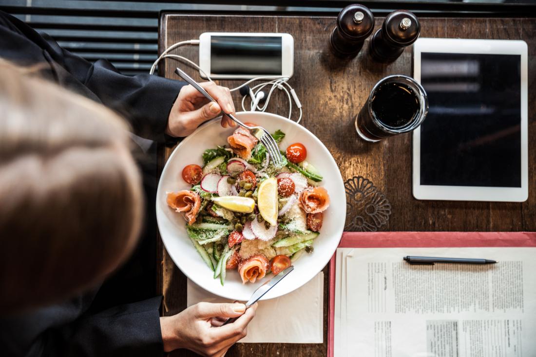Person eating a salad with phone, tablet, and folder on table.