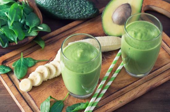 Green smoothies in glasses on wooden chopping board, with spinach leaves, banana, and avocado.