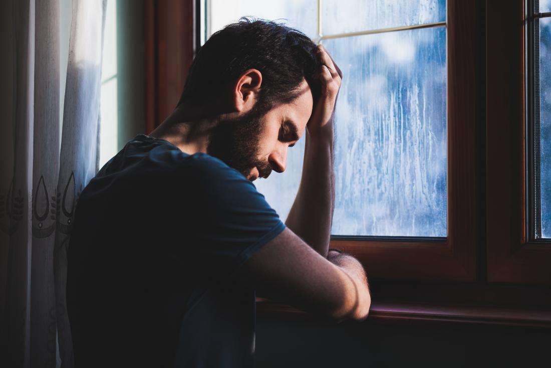 Man at window holding head, looking sad and pensive.