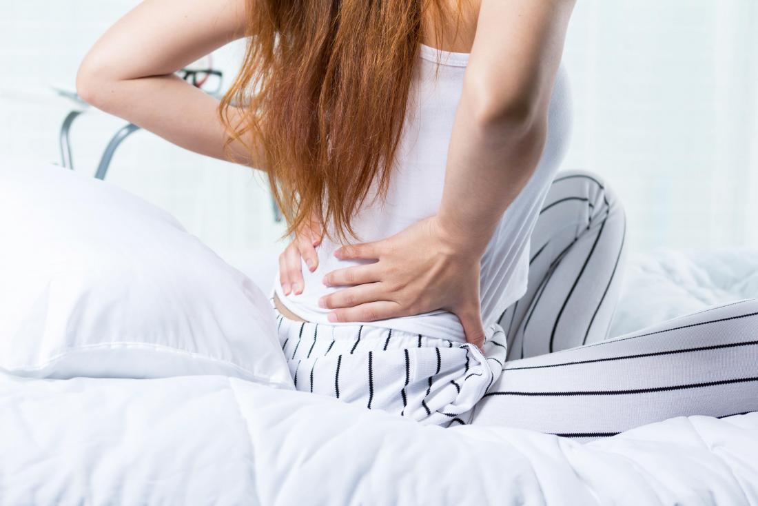 Woman sitting on bed holding lower back in pain trying to sleep.