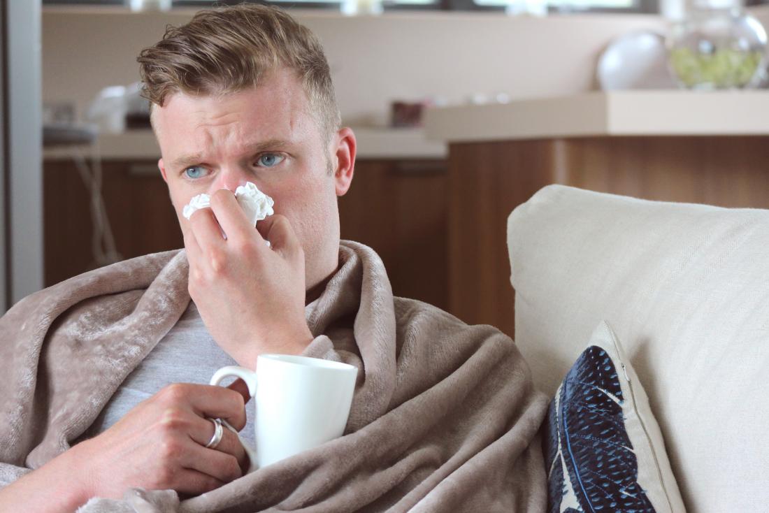 man with runny nose drinking hot drink at home under blanket on sofa