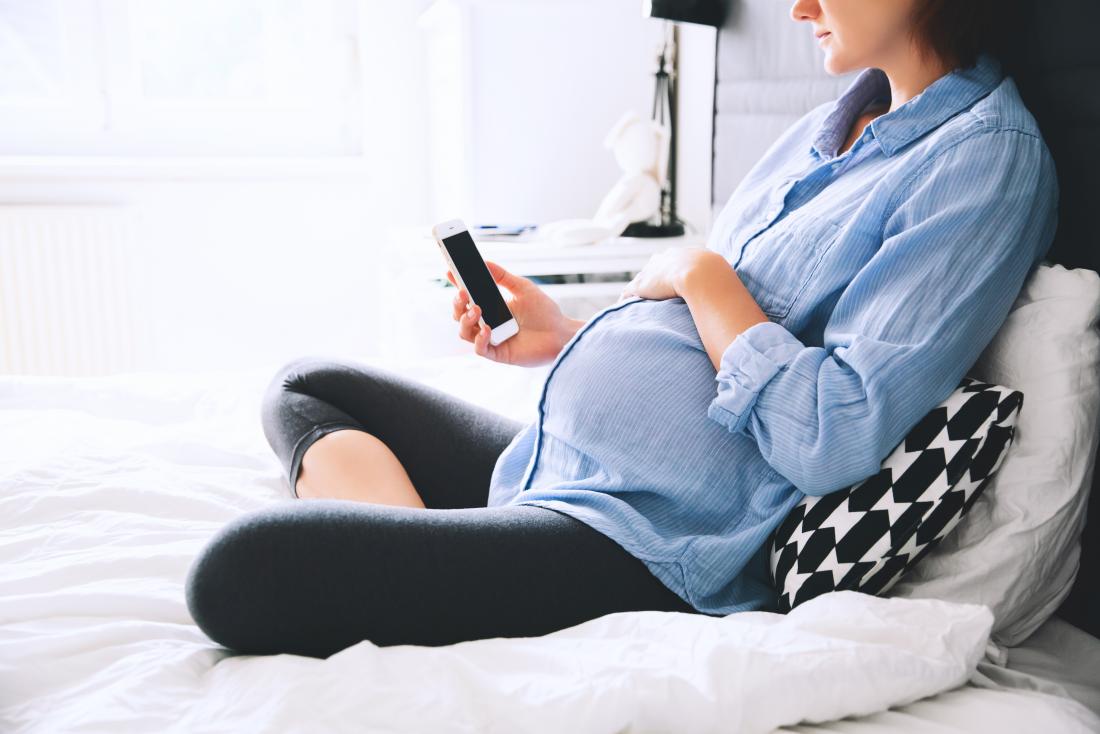 pregnant woman sitting on bed using smartphone