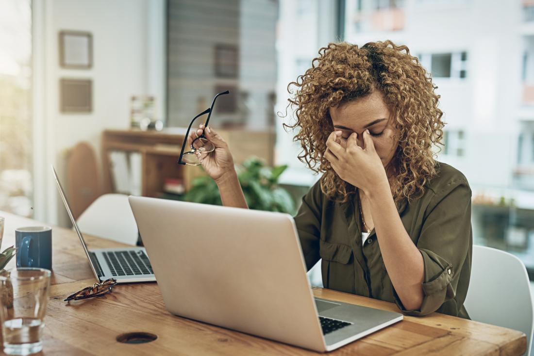 Stressed woman working in home office with two laptops rubbing bridge of nose because of tension headache.