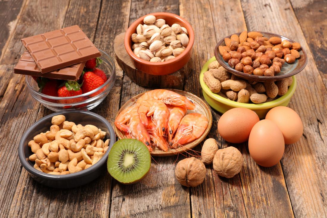 Allergy foods laid out on wooden table, including cashew nuts, pecans, hazelnuts, almonds. chocolate, strawberries, shellfish, walnuts, eggs, and kiwi.