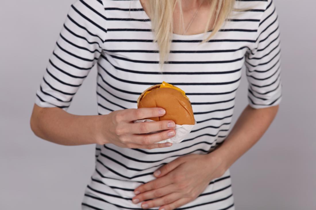Woman with stomach pain after eating holding a burger and a hand in front of her tummy.