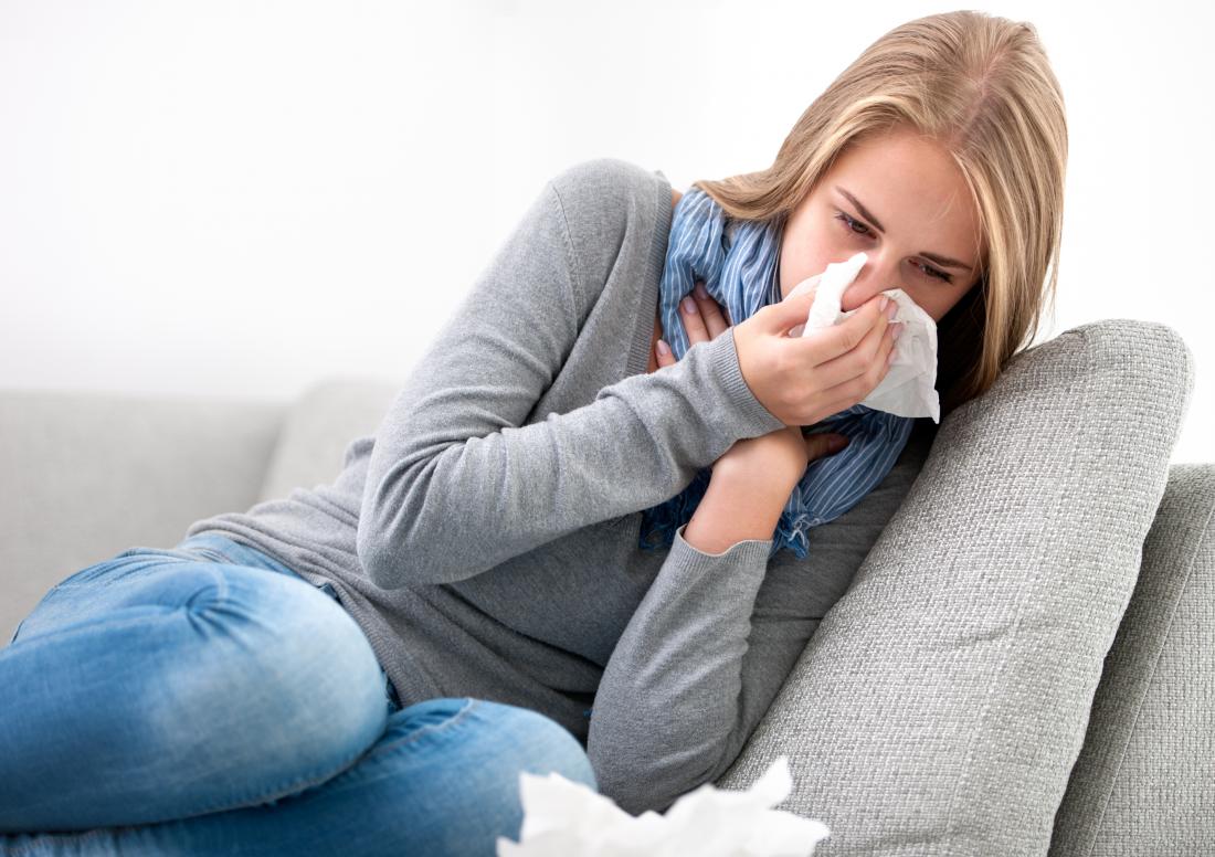 Woman blowing nose on tissues while sitting on sofa unwell.