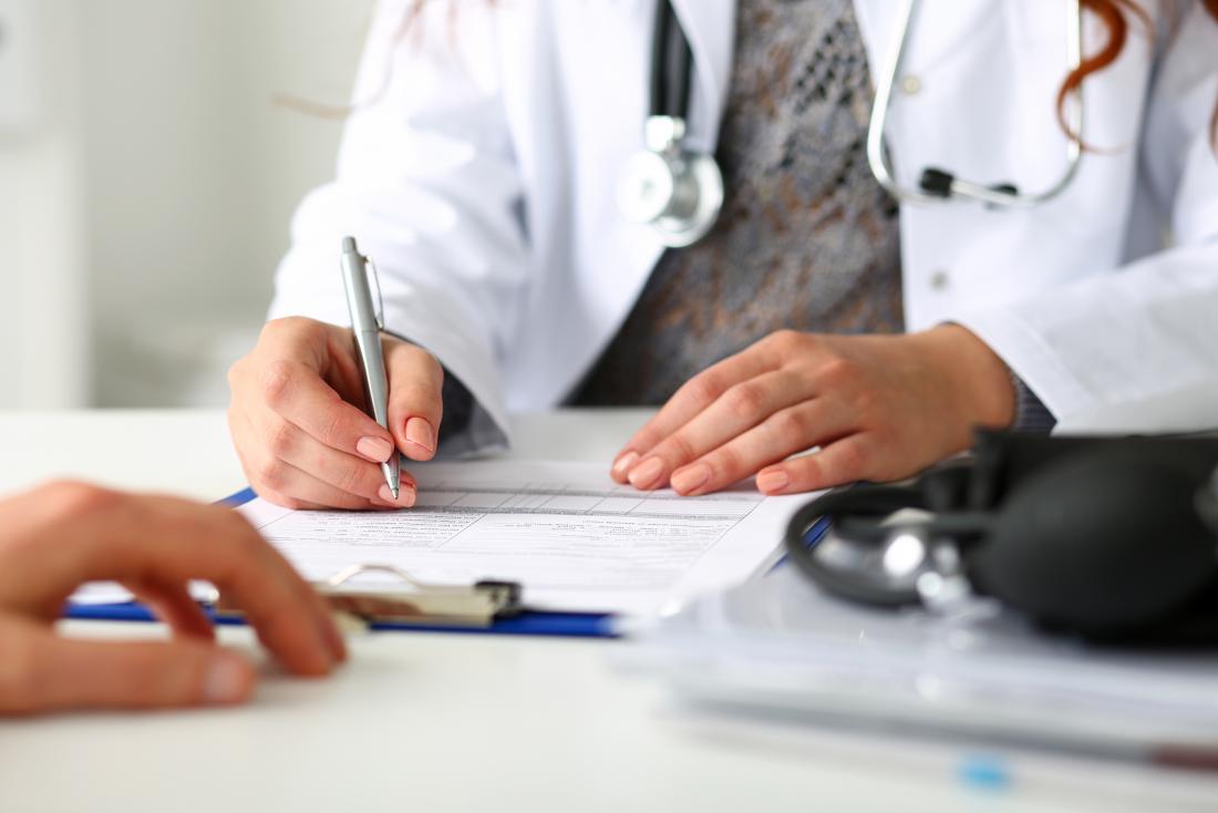 Female doctor writingo n clipboard at desk with patient in foreground.