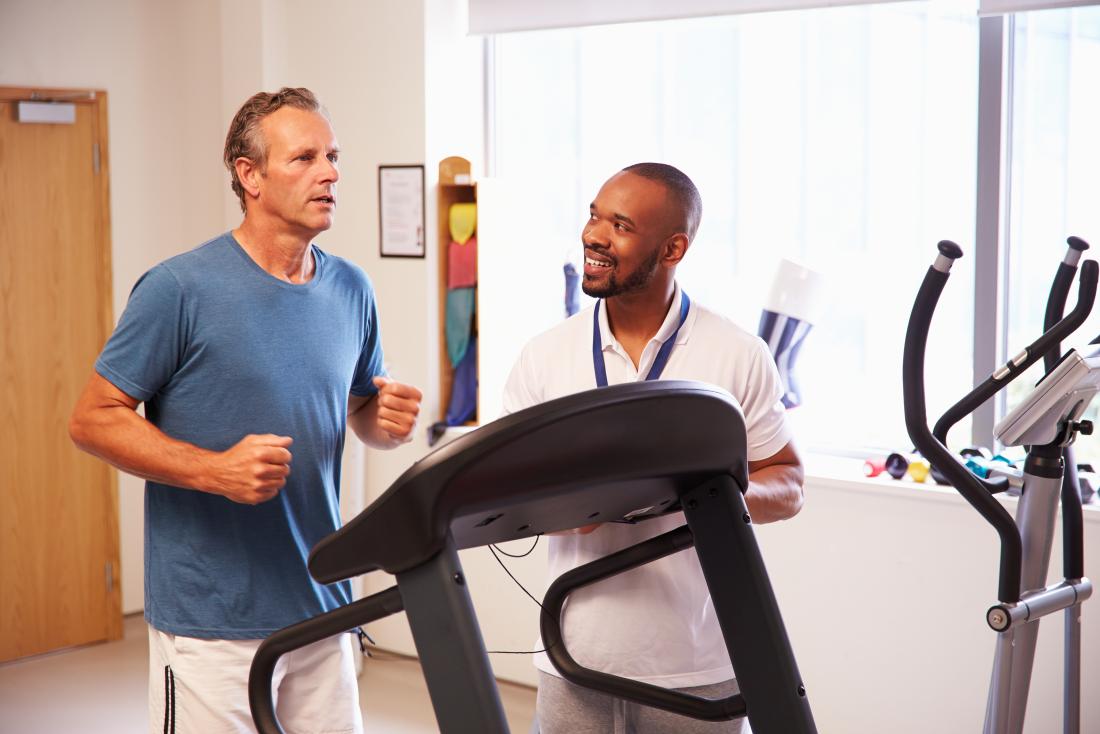 Man on treadmill in physical therapy clinic with trainer monitoring.