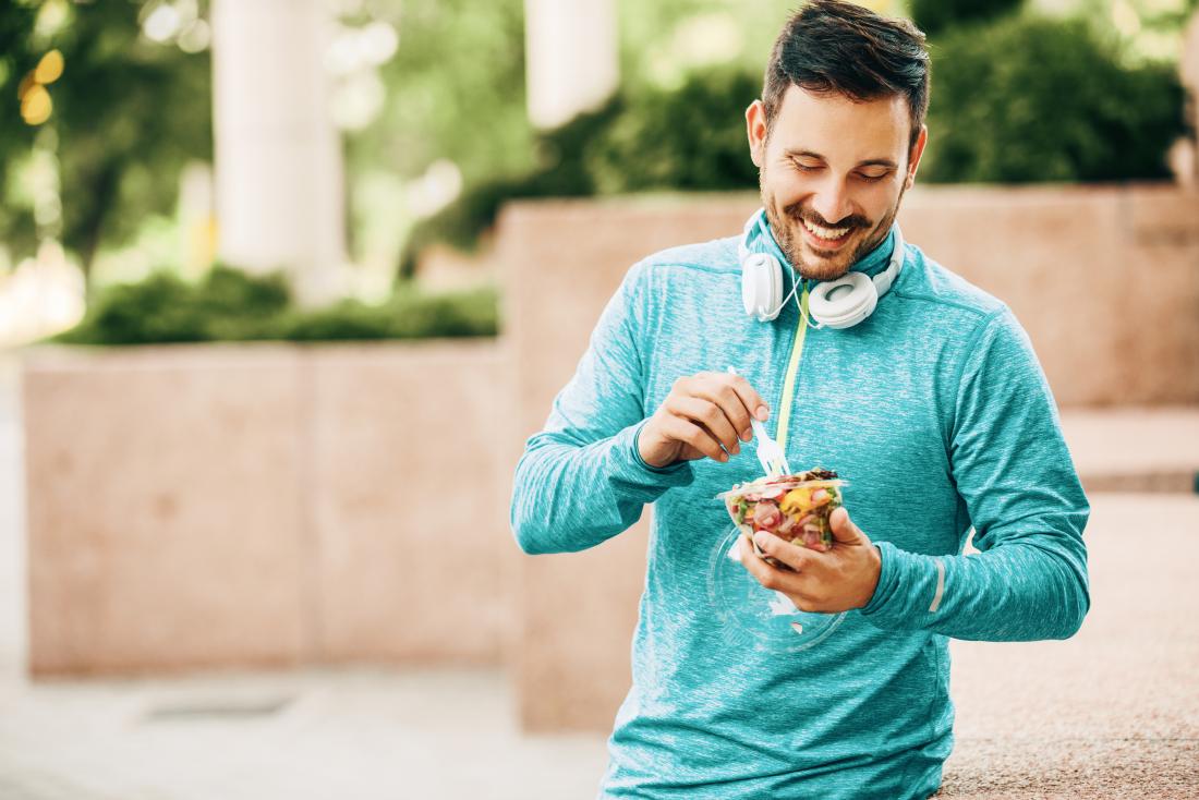 Man in running gear outdoors eating from takeaway pot of pasta.