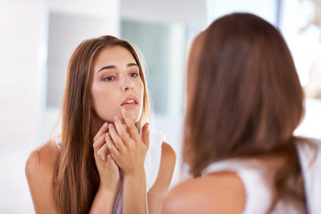 Woman touching lip because of pimple of cold sore, looking in mirror