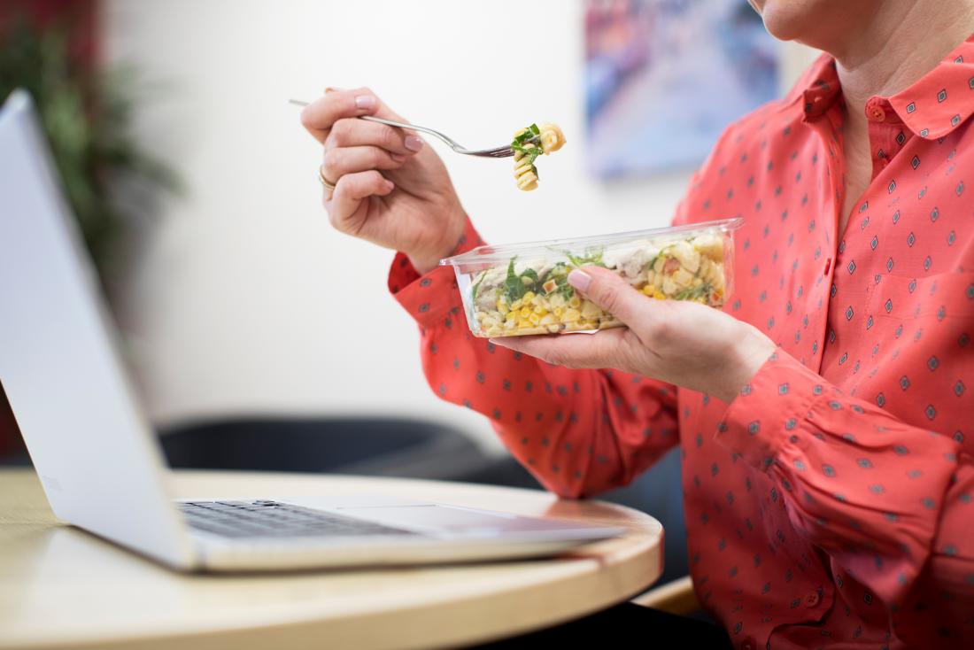 Person eating pasta lunch at desk in office in front of laptop