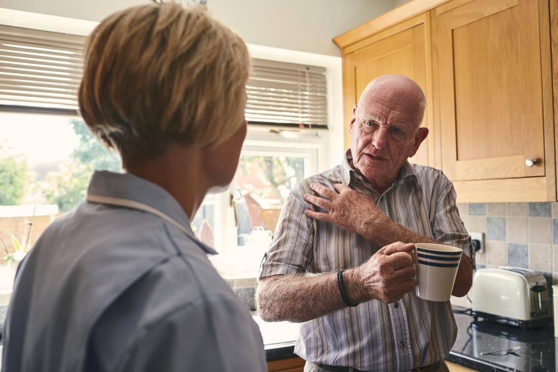 Senior man in kitchen holding shoulder because of pain showing home care nurse