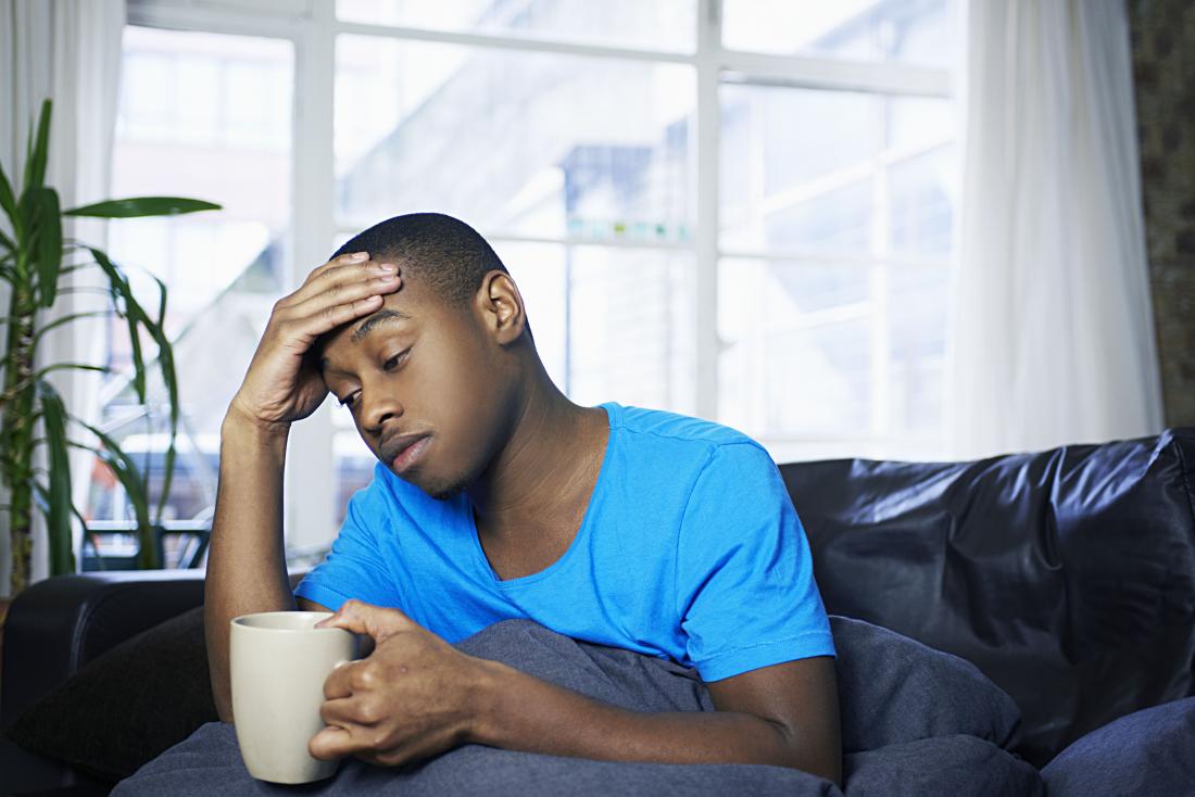 Man holding head looking ill with mug under blanket
