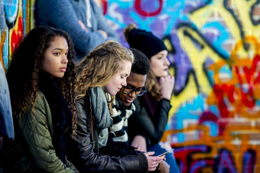 Group of teenagers sit in a row in front of a wall of graffiti.