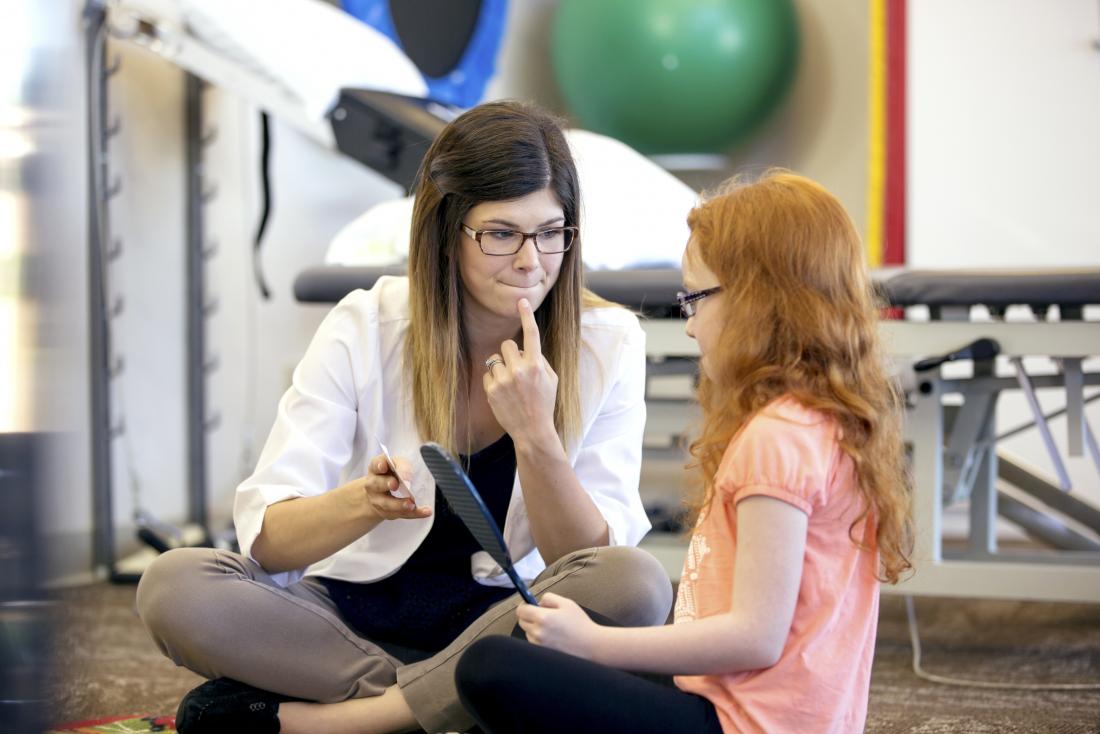 speech therapy being conducted by doctor sitting with young girl who is holding a mirror