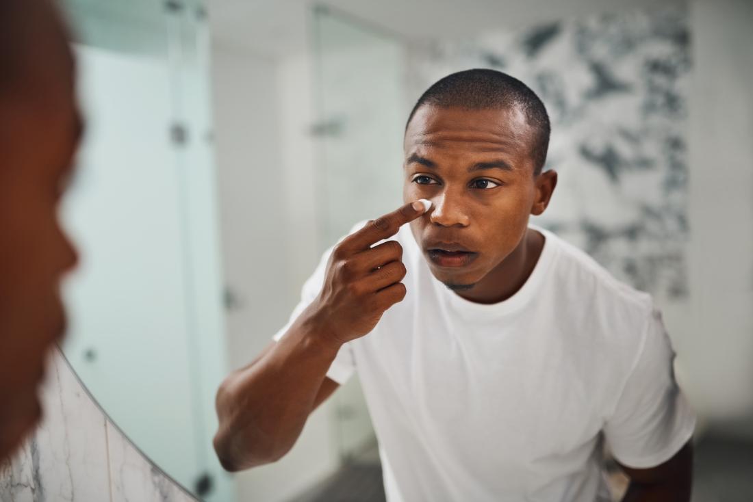 black or african american man looking in bathroom mirror applying cream or lotion to skin under eyes